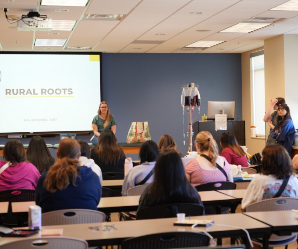 High school students sit in a classroom facing a screen with "Rural Roots" written on it. An RVU student faces the group.