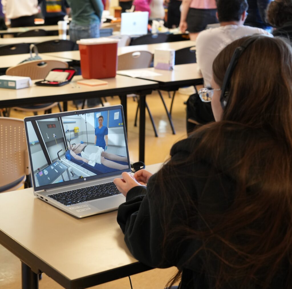 A high school sits at a desk looking at a laptop where a virtual reality simulation of a hospital room is being shown